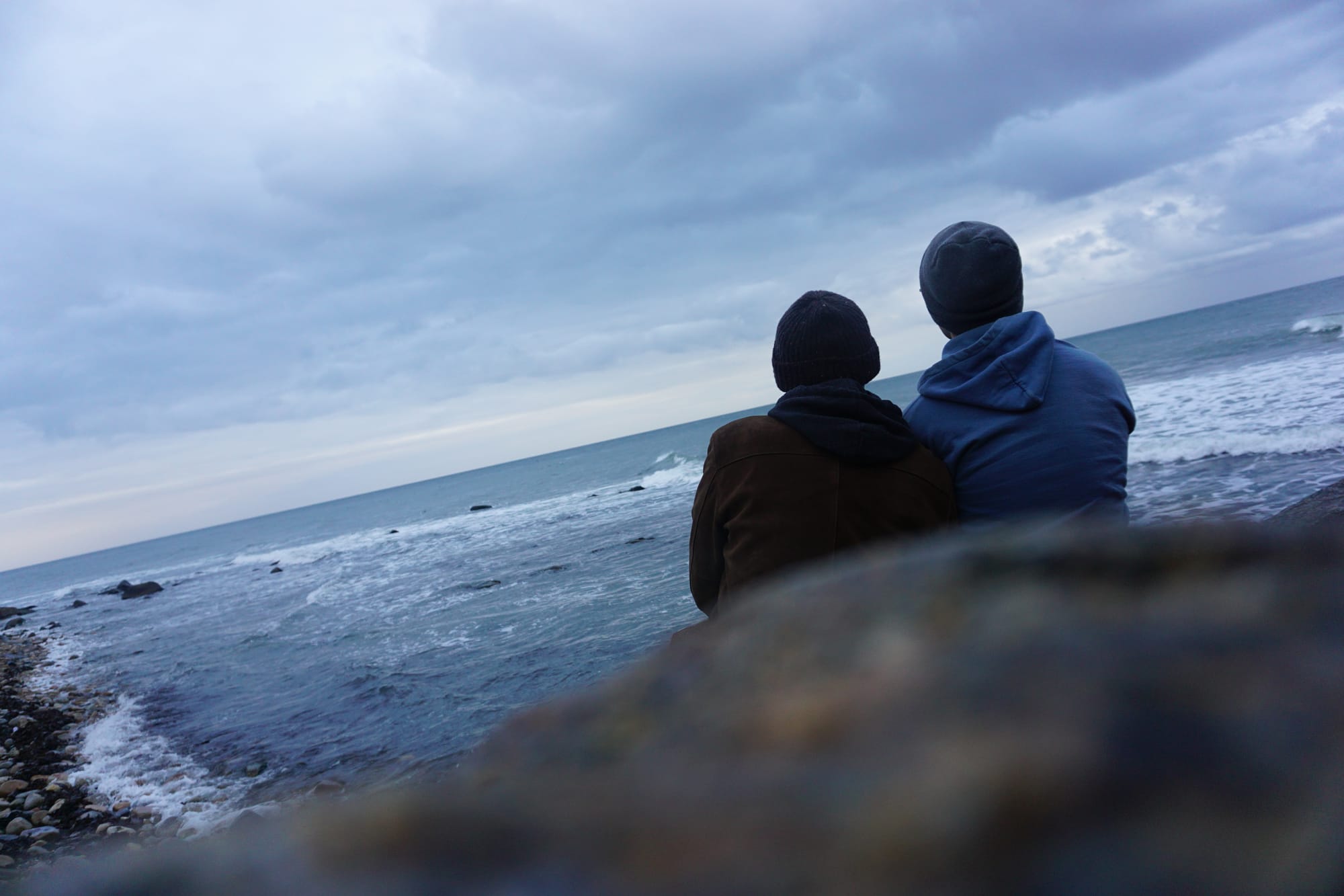 Two people sit together on a rocky Montauk beach in 2020, facing the ocean under a cloudy sky, wrapped in warm clothing.